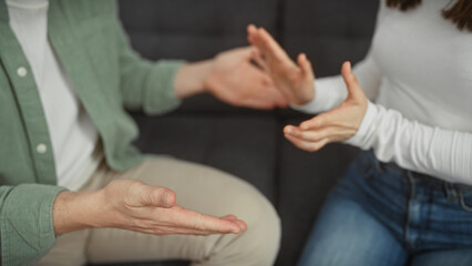 A man and woman engage in a discussion with expressive hand gestures in the living room of a home. - obrazy, fototapety, plakaty