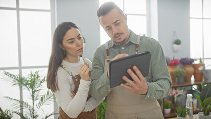 Man and woman in aprons reviewing a tablet together at a flower shop with plant arrangements in the background