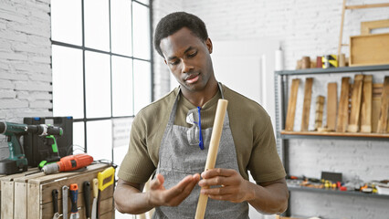 A focused african american man inspects a wooden piece in a well-equipped carpentry workshop.