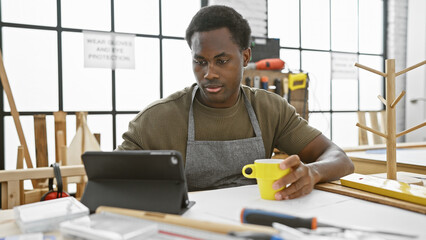 Focused african man using tablet in sunlit carpentry studio, holding coffee, tools around.