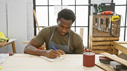 African american man writing in a notebook at a carpentry workshop indoors