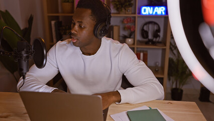 Adult african american man podcasting at night in his home office, using a microphone and laptop.