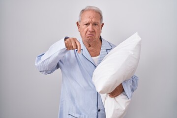 Senior man with grey hair wearing pijama hugging pillow pointing down looking sad and upset,...