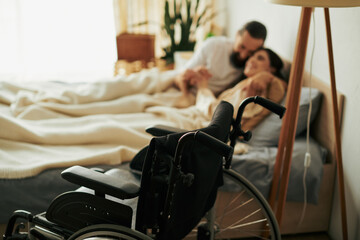 focus on modern wheelchair in front of blurred loving couple lying in bed together while at home