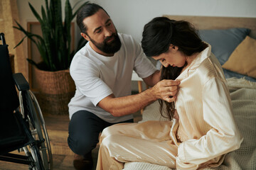 bearded loving man in homewear helping his disabled wife to change into pajama in bedroom at home