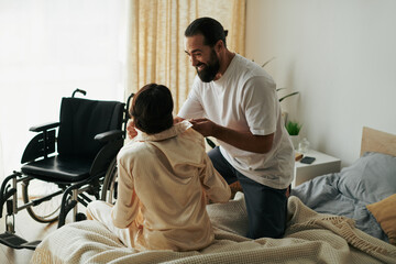 handsome loving man in homewear helping his disabled wife to change into pajama in bedroom at home