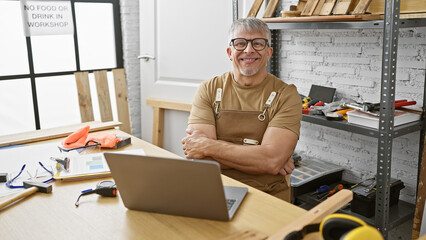 Smiling grey-haired man in apron with crossed arms standing in a carpentry workshop with tools and...