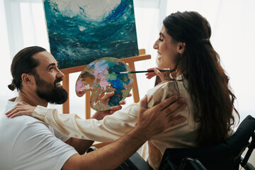 appealing woman with mobility disability painting on easel next to her cheerful bearded husband - obrazy, fototapety, plakaty