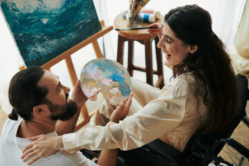 appealing woman with mobility disability painting on easel next to her cheerful bearded husband