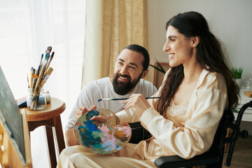 positive husband helping his inclusive beautiful wife on wheelchair to paint on easel while at home