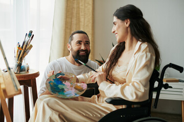 positive husband helping his inclusive beautiful wife on wheelchair to paint on easel while at home - obrazy, fototapety, plakaty