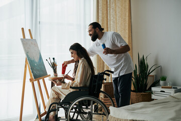 caring cheerful couple of bearded man and disabled woman painting on easel together at home - obrazy, fototapety, plakaty