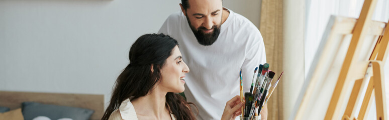 joyous husband helping his inclusive beautiful wife on wheelchair to paint on easel, banner - obrazy, fototapety, plakaty