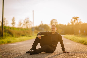 Valmiera, Latvia - August 17, 2024 - A man sits on a country road with a laptop on his knees,...