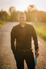 Obraz premium Valmiera, Latvia - August 17, 2024 - A man stands on a country road at sunset, holding a folder, with a pleasant smile on his face.