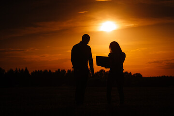 Fototapeta na wymiar Valmiera, Latvia - August 17, 2024 - Silhouettes of a man and a woman with a laptop in a field during a vibrant sunset.
