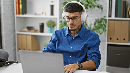 Handsome young latin man jamming to music while working on laptop in elegant office, a tattooed...