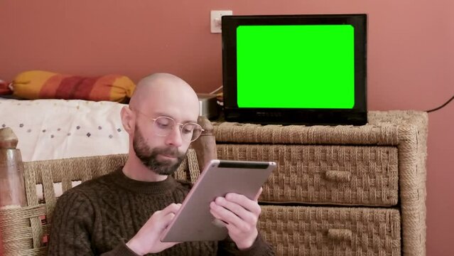 young man with beard uses tablet, old TV 80s with green screen mockup, exotic Eastern woven furniture, capturing authentic moment downshifting life, work-life balance