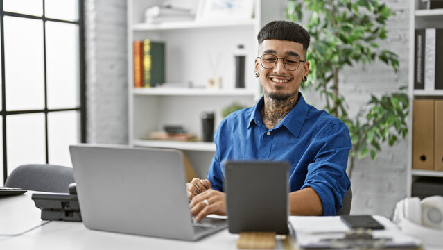 Confident young latin man, smiling happily while working online using his laptop and touchpad at a professional business office