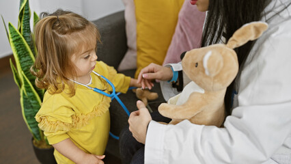 A young girl in a yellow dress plays doctor with a plush toy as a woman assists her in a cozy home...