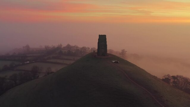 Misty sunrise aerial of Glastonbury Tor, colourful sky with countryside in the background, Somerset, England