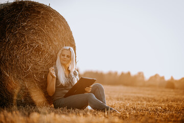 Valmiera, Latvia - August 17, 2024 - A woman is sitting in a field at sunset, holding a tablet and...