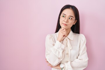 Young chinese woman standing over pink background with hand on chin thinking about question,...