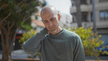 A pensive bald man with a beard touches his neck in discomfort while standing outdoors in a green...