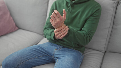 Young bald man in casual attire experiencing wrist pain at home, sitting on a gray sofa.