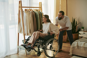handsome caring man helping his inclusive wife on wheelchair to get dressed while in bedroom - obrazy, fototapety, plakaty