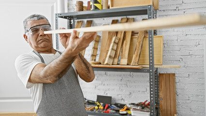 Mature man in safety glasses meticulously examines a wooden plank in a well-organized carpentry...