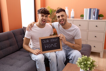 Homosexual gay couple holding blackboard with first home text winking looking at the camera with sexy expression, cheerful and happy face. - obrazy, fototapety, plakaty
