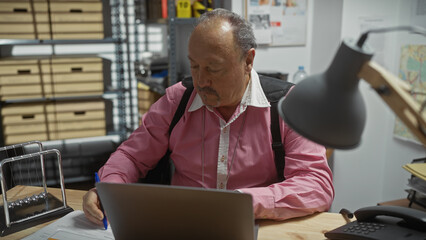 Mature man working indoors at a cluttered office desk with paperwork, laptop, and phone, showing...