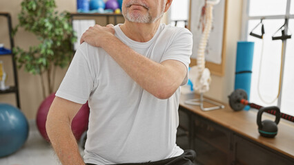 Senior man experiencing shoulder pain in a physiotherapy clinic with rehabilitation equipment...