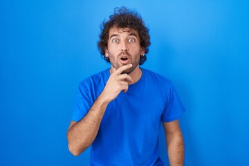 Hispanic young man standing over blue background looking fascinated with disbelief, surprise and...