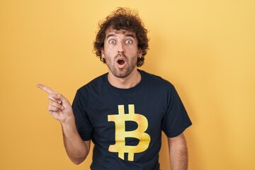 Hispanic young man wearing bitcoin t shirt surprised pointing with finger to the side, open mouth...
