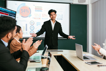 A cheerful and confident Asian businessman stands, presenting bar charts data from the projector screen to his office colleagues. Asian businessman leader role at the meeting. - 785326243