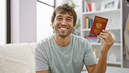 Handsome blond young man, a confident patriot, joyfully smiling with his polish passport, casually lounging on his living room sofa, ready for european holiday adventures!