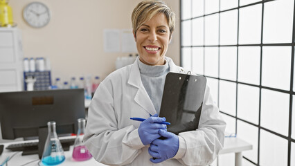 A confident hispanic woman with short hair smiling in a laboratory, holding a clipboard, wearing a...