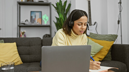 Hispanic woman studying in a modern living room with a laptop and headphones, writing notes.