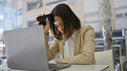 A stressed young hispanic woman at a desk with a laptop holding her phone to her temple in a beige office environment.