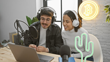 A man and woman with headphones collaborate in a modern podcast studio with a laptop and bitcoin...