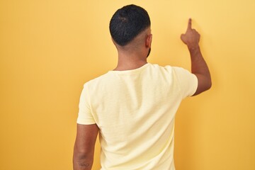 Young hispanic man standing over yellow background posing backwards pointing ahead with finger hand