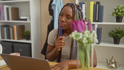 Thoughtful african woman with braids pondering indoors at a living room table, near laptop and...