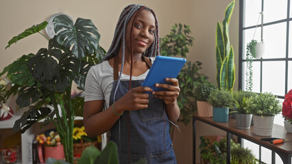African american woman with braids using a tablet in a flower shop with green plants in the...
