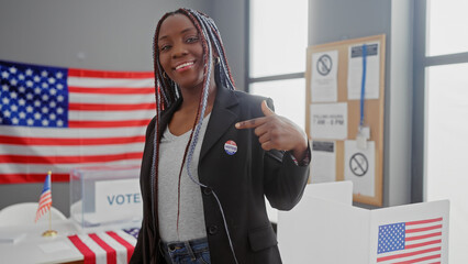 African american woman with braids pointing at her 'i voted' sticker in a college electoral center...