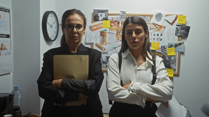 Two serious women detectives stand with crossed arms in a police department investigation room, surrounded by evidence. - obrazy, fototapety, plakaty