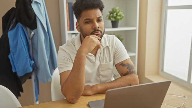 Pensive african american man with tattoo at home using laptop in a living room.