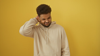 A confused african man scratching his head against an isolated yellow background, portraying a...