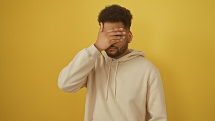 A distressed man covers his face with hand against a yellow background, embodying worry, fear or...